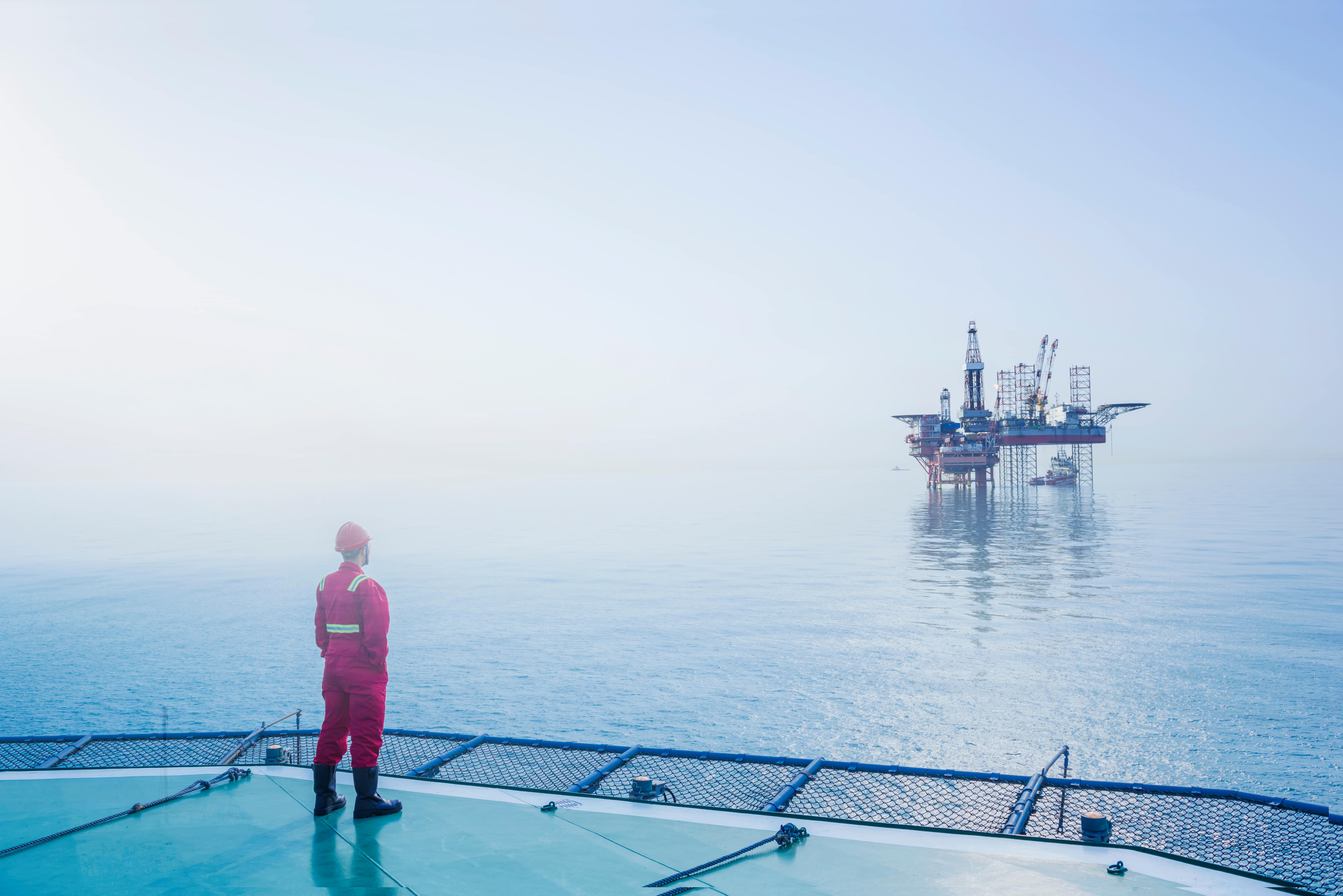 An offshore drill worker overlooking an oil rig in a vast, blue ocean.