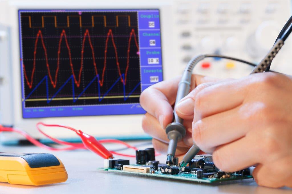 Engineer testing an electronic circuit board using oscilloscope probes while monitoring signal waveforms on a digital oscilloscope.