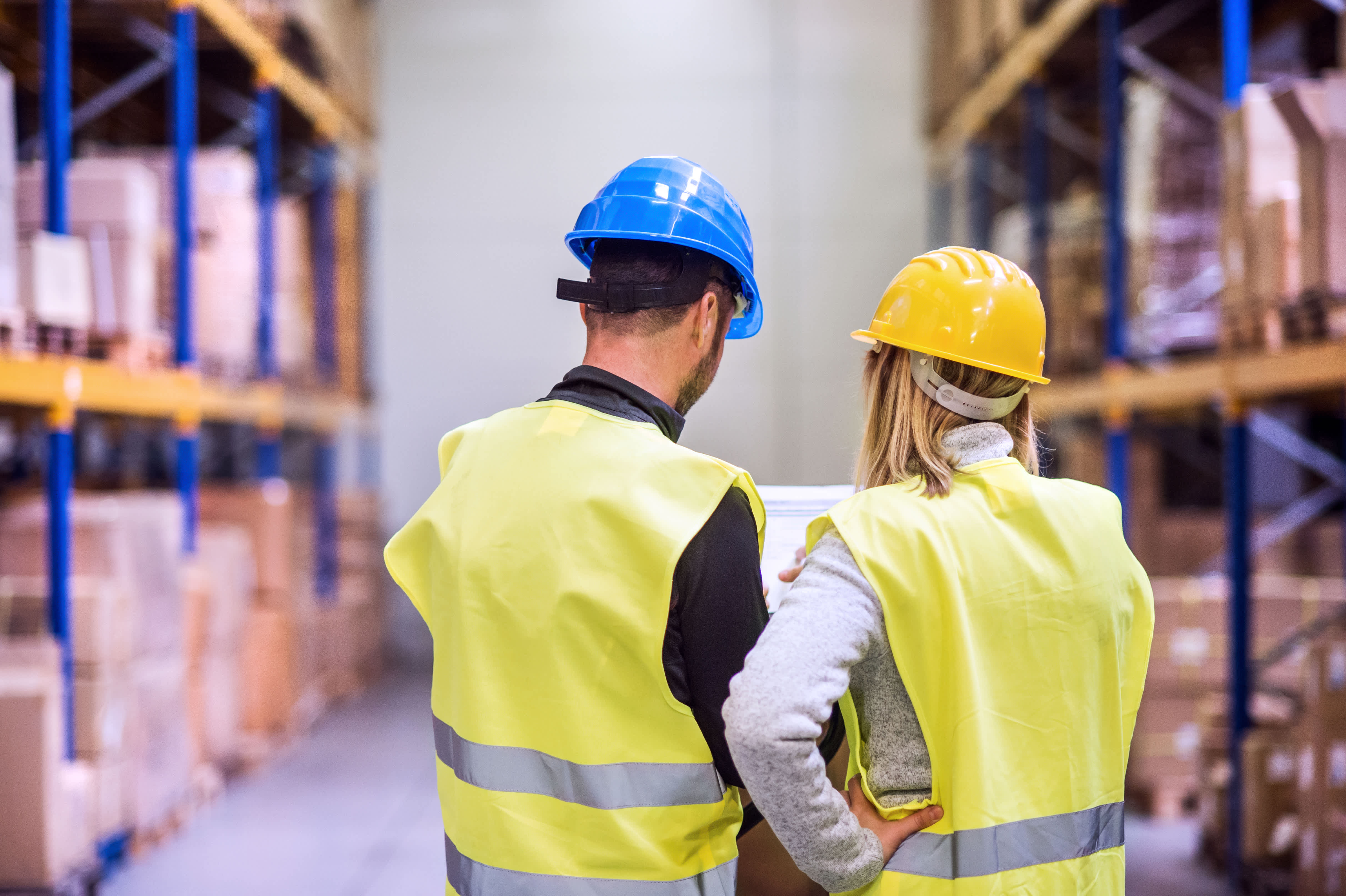 Two workers in hi-vis standing in between two large shelves in a warehouse, looking at a clipboard.