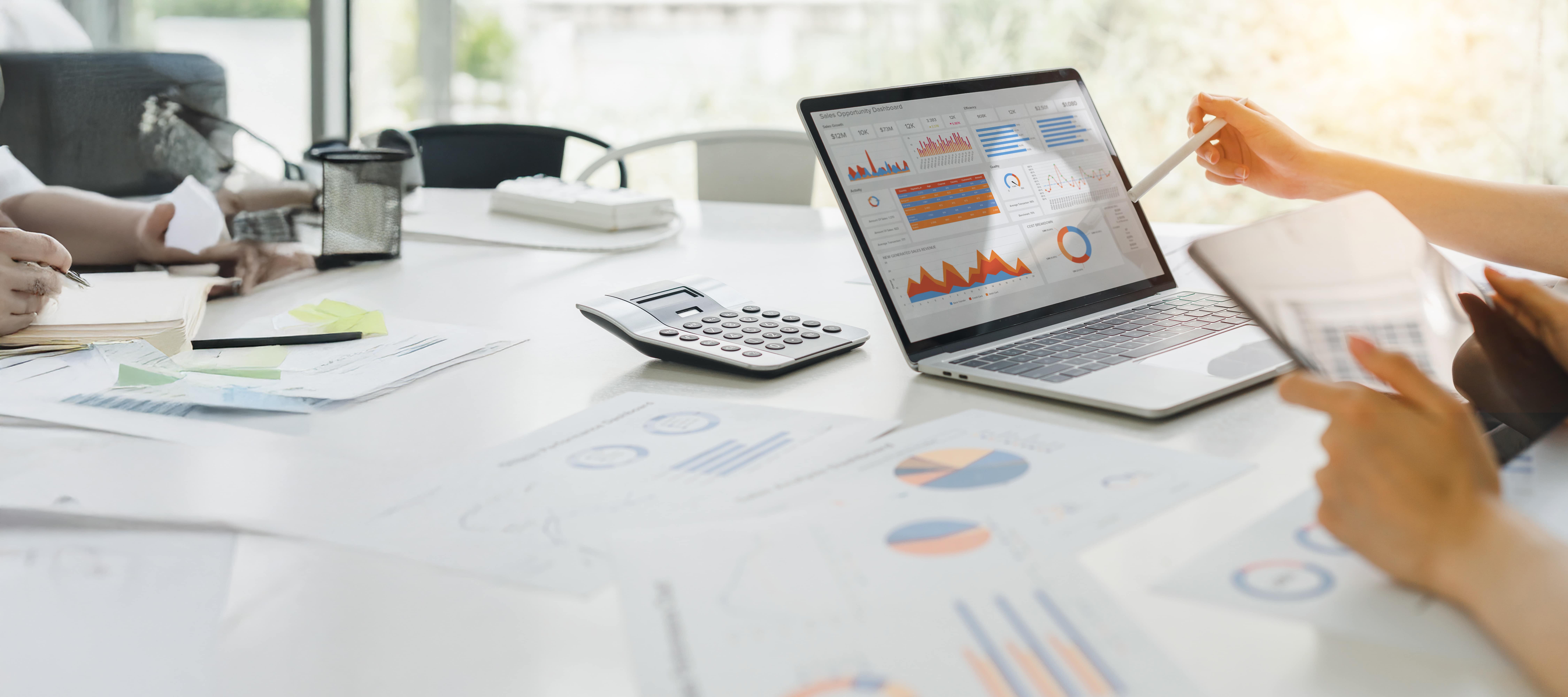 A person in a meeting, pointing towards a laptop showing pie charts and graphs of sales information