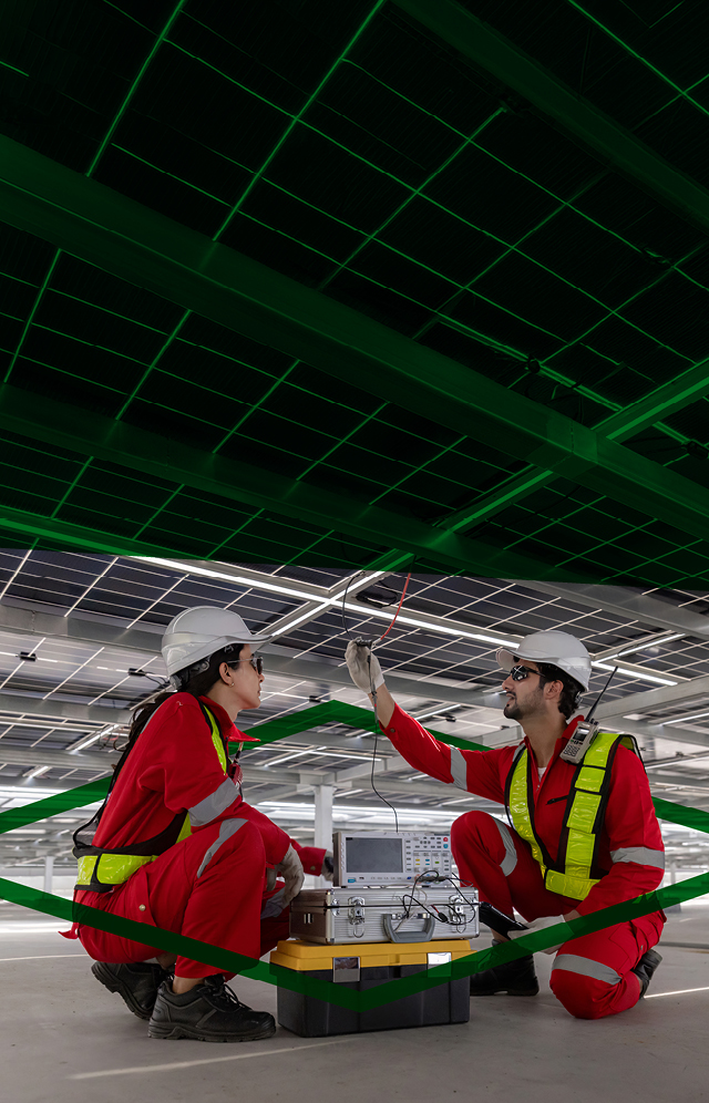 Two workers wearing hi-vis clothing and safety helmets working with electrical equipment in a large warehouse.