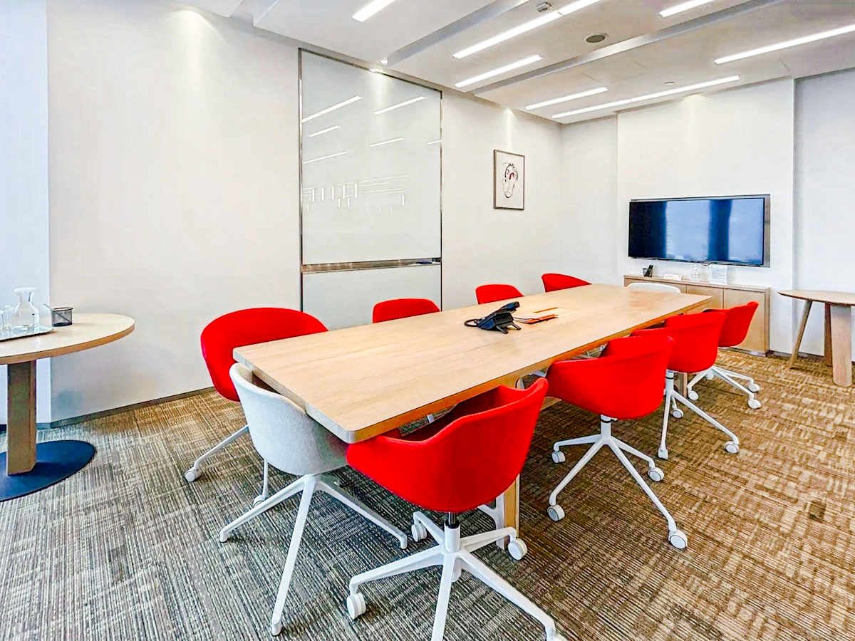 Modern coworking space with bright red chairs, sleek meeting table, and wall-mounted screen.