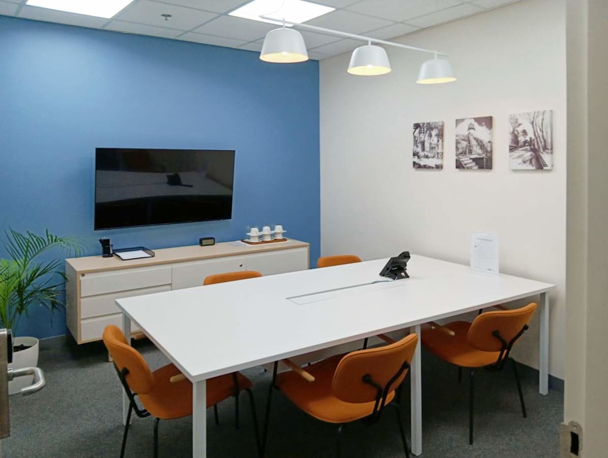 Bright, modern meeting room with a sleek white table, orange chairs, and wall-mounted TV.