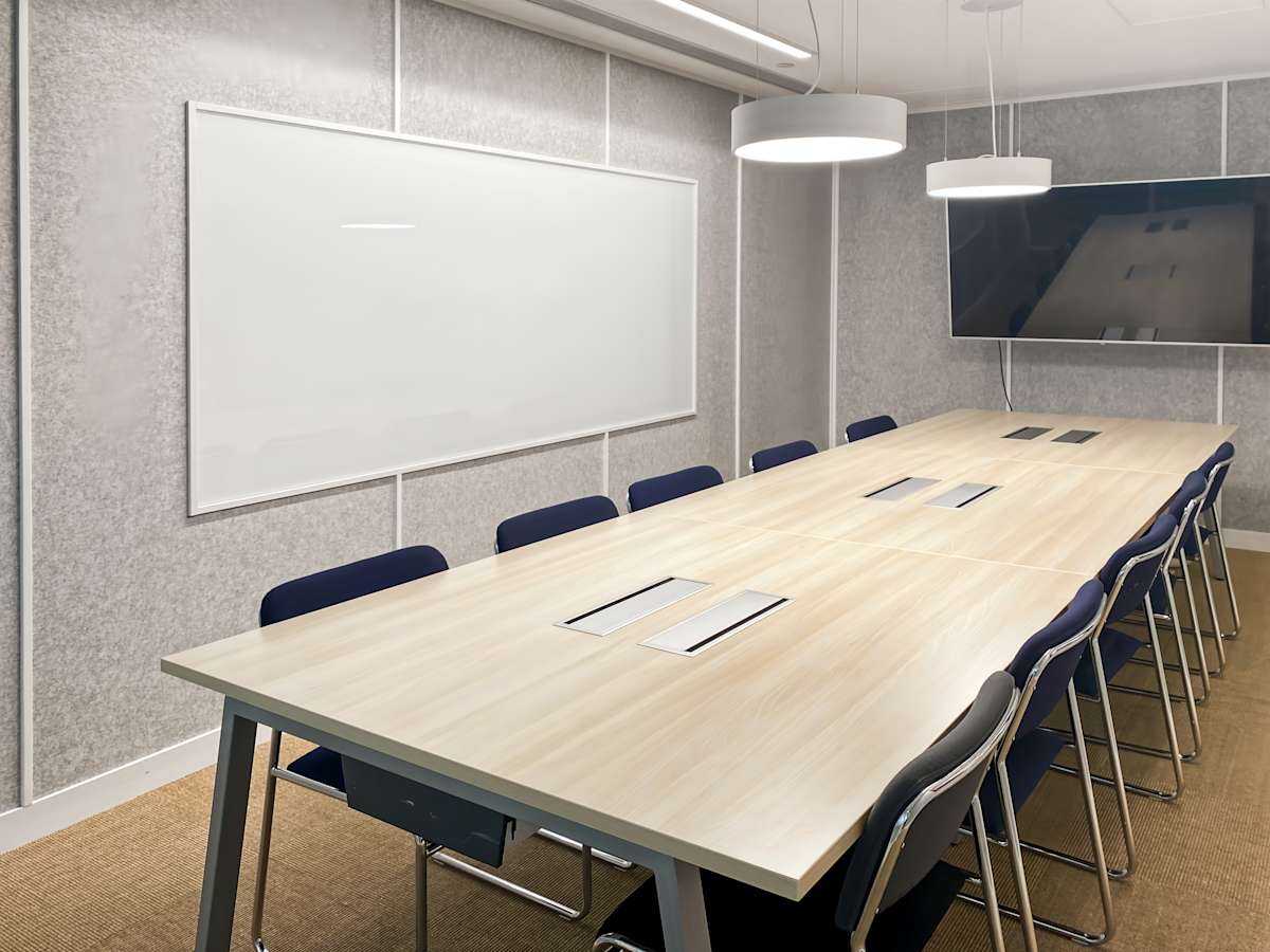 Modern meeting room with a long table, cushioned chairs, whiteboard, and mounted screen.
