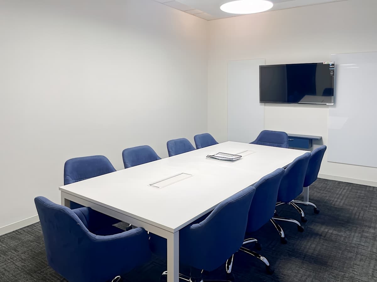 Modern meeting room with a sleek white table, comfortable blue chairs, and a wall-mounted screen.