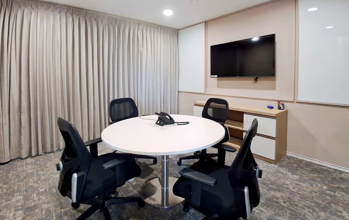 Modern meeting room with round table, ergonomic chairs, wall-mounted TV, and ample lighting.
