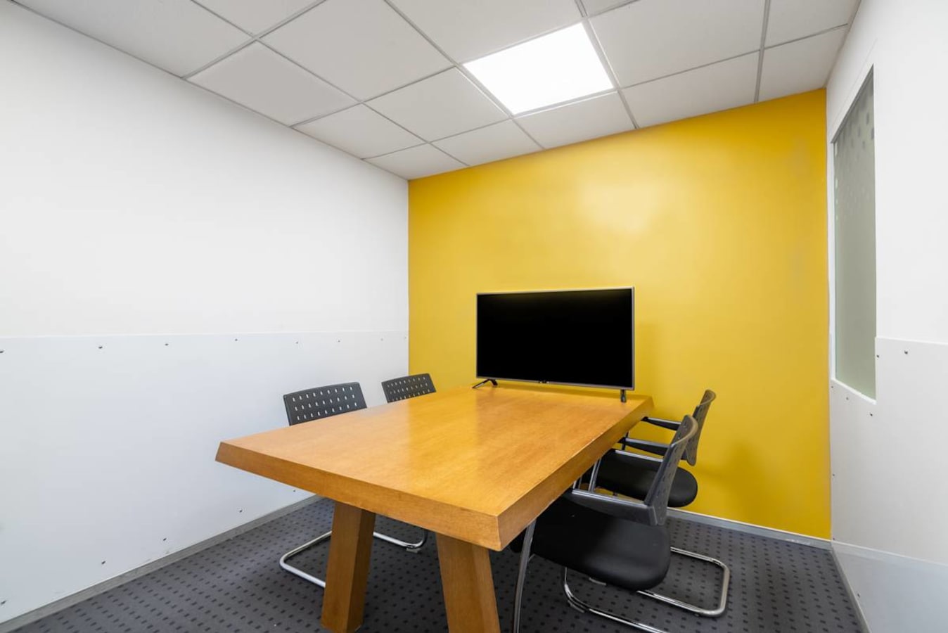 Bright, modern meeting room with a wooden table, sleek chairs, and a wall-mounted screen.
