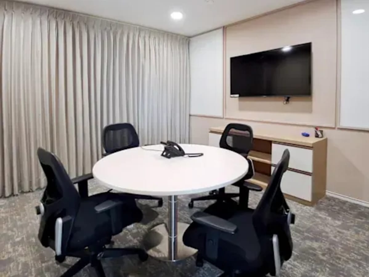 Modern meeting room with round table, sleek chairs, wall-mounted TV, and natural lighting.