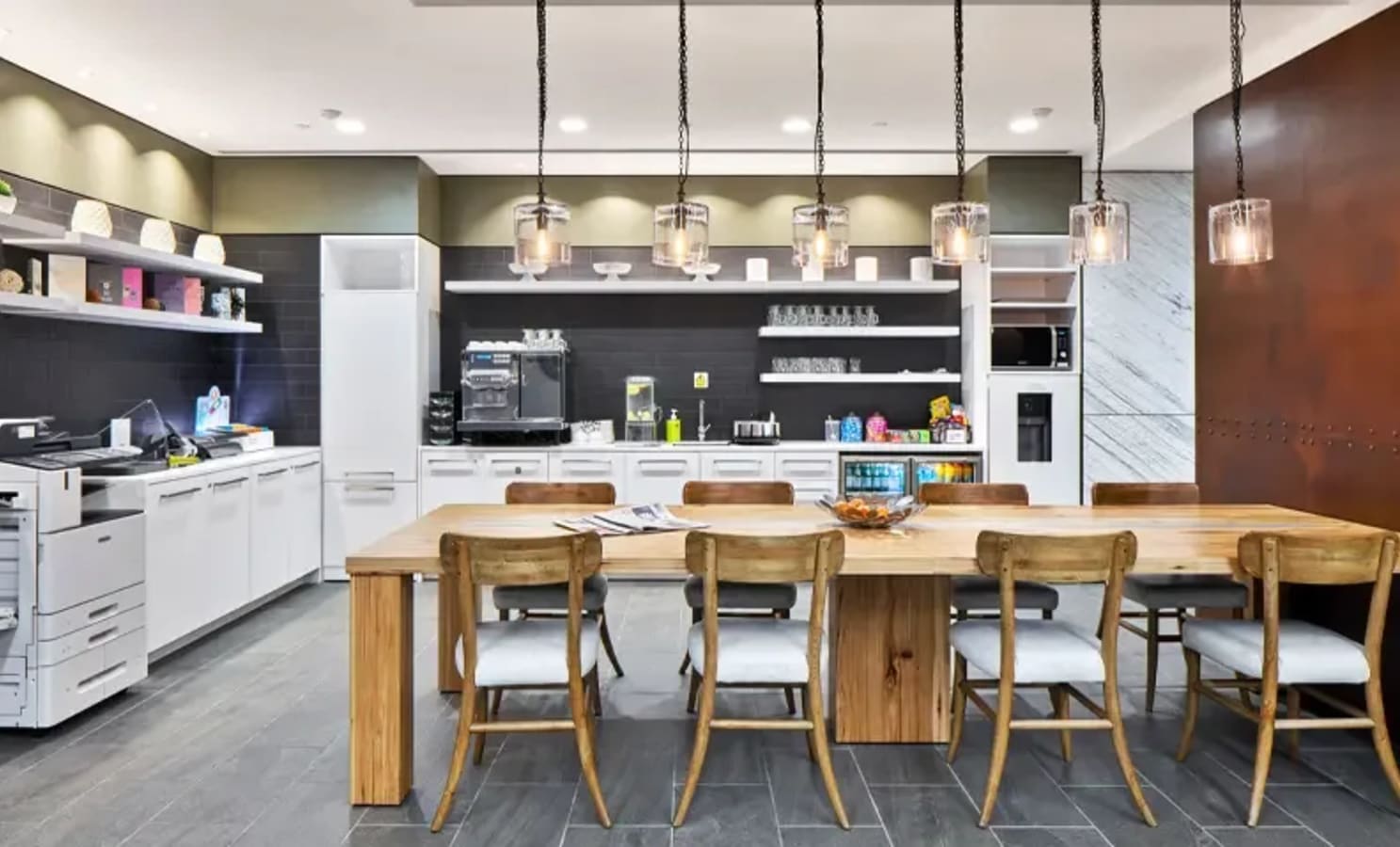 Modern coworking kitchen with communal table, hanging lights, and full amenities. Ideal for collaboration.