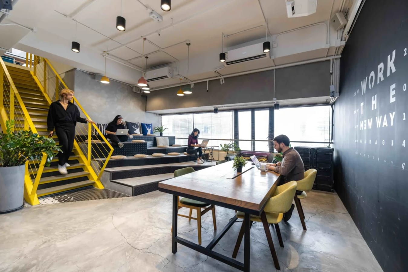 Modern coworking space with open seating, vibrant yellow stairs, and abundant natural light.