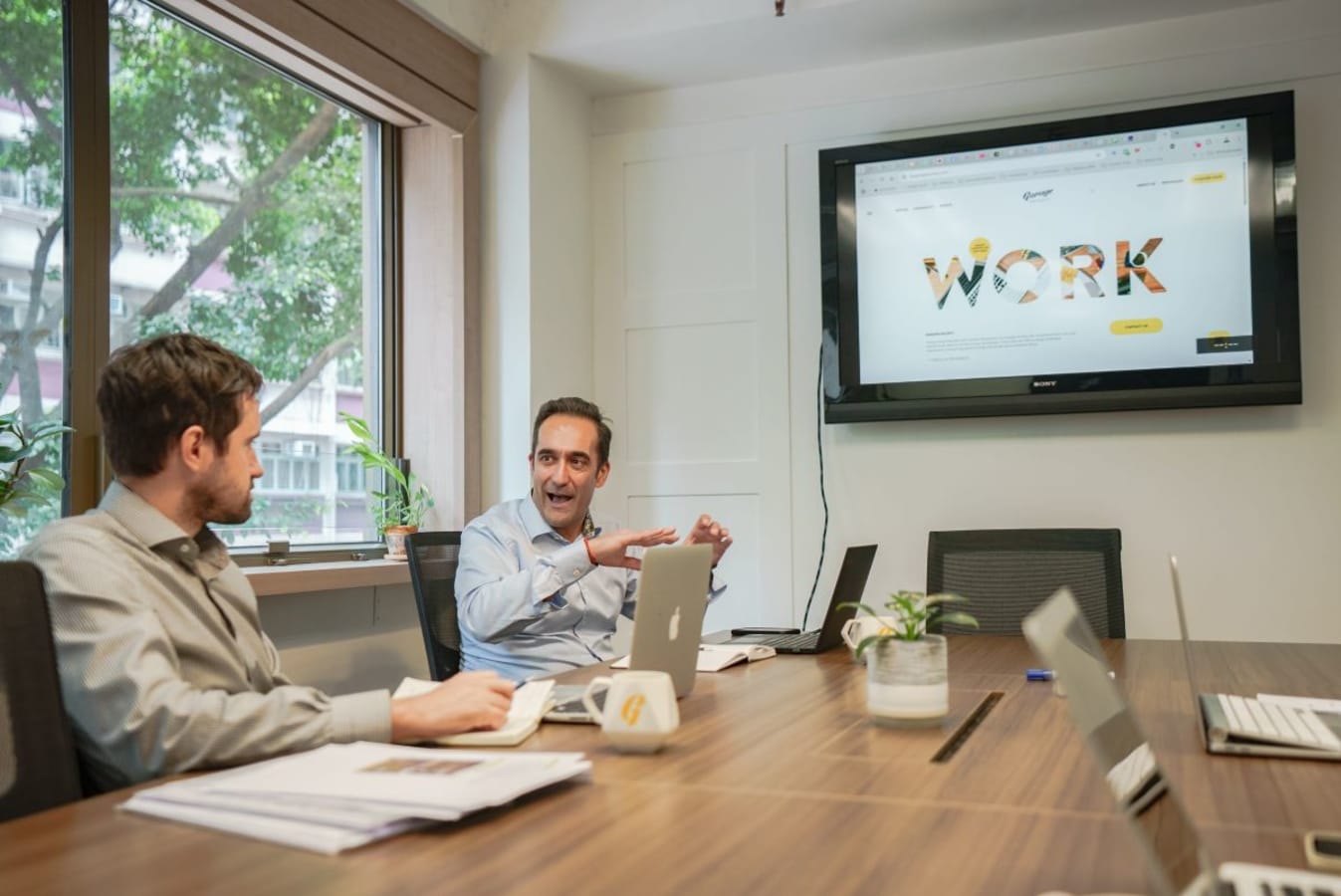 Modern coworking space with natural light, conference table, and a large screen for presentations.