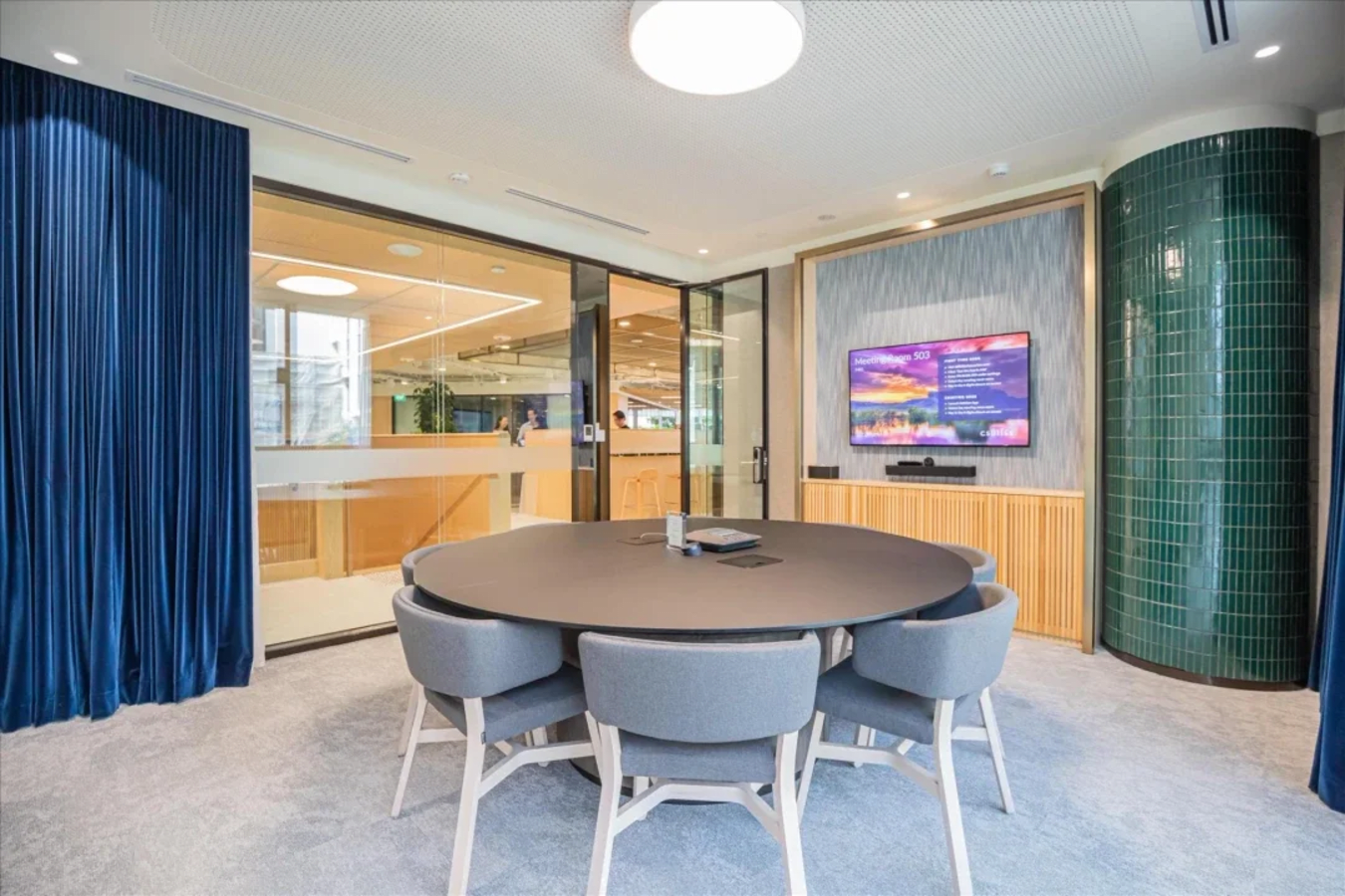 Modern meeting area with round table, plush seating, smart TV, and ample natural light.