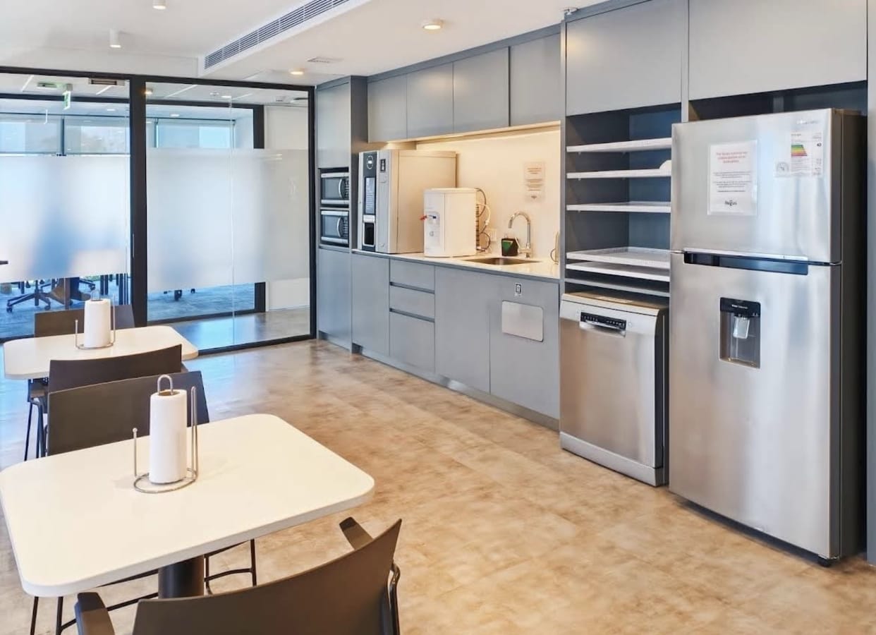 Modern kitchenette with seating, stainless appliances, and ample storage in a bright coworking space.