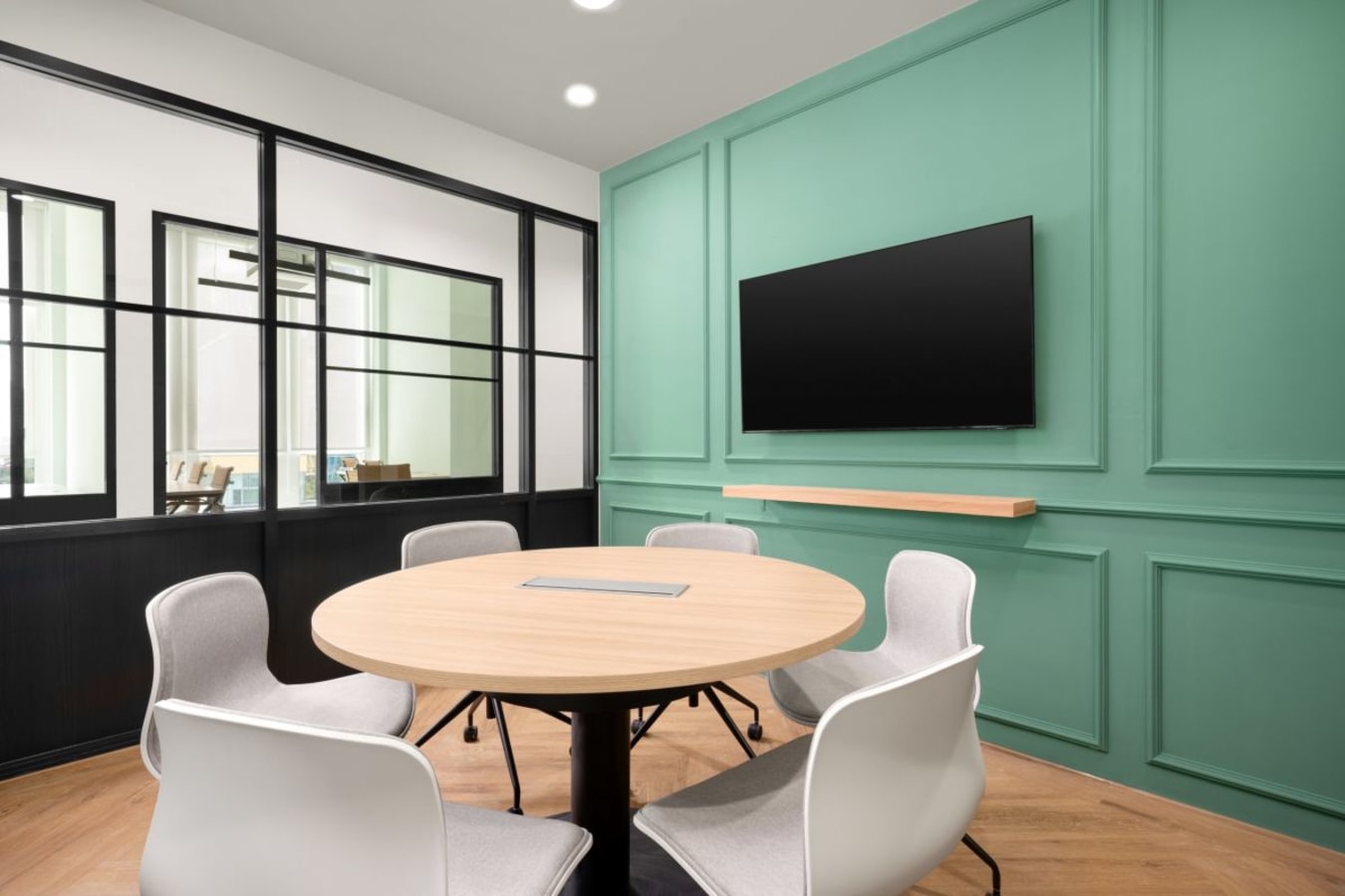 Modern meeting room with a round table, ergonomic chairs, and a wall-mounted screen.