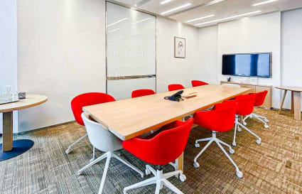 Modern coworking space with bright red chairs, sleek meeting table, and wall-mounted screen.