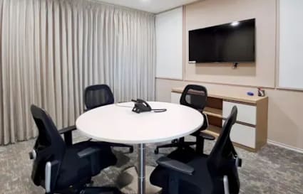 Modern meeting room with round table, sleek chairs, wall-mounted TV, and natural lighting.