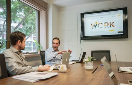 Modern coworking space with natural light, conference table, and a large screen for presentations.