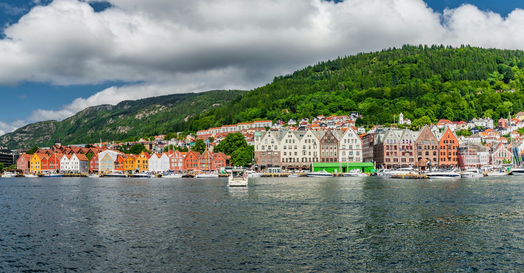 Et panoramabilde av Bryggen i Bergen, Vestland med sine ikoniske fargerike trehus langs havnebassenget, omgitt av grønne fjellsider under en skyet sommerhimmel.