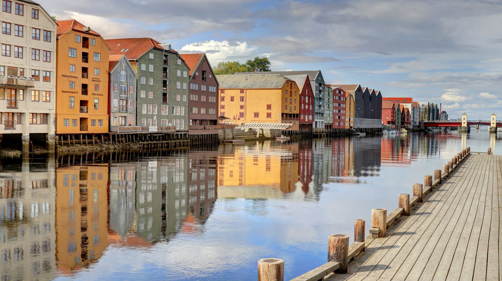 En rekke fargerike trebygninger langs Nidelva i Trondheim, kjent som de gamle bryggene, reflekteres tydelig i det stille vannet.