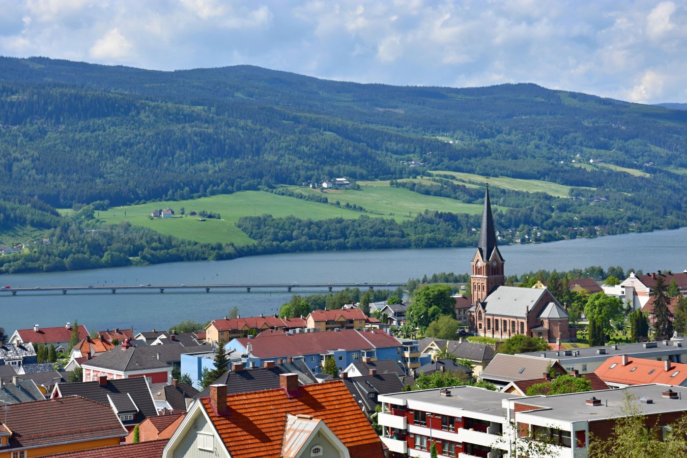 Panoramautsikt over Lillehammer. Byen er omgitt av grønne åser, med Mjøsa og en lang bro i bakgrunnen under en blå himmel med lette skyer.