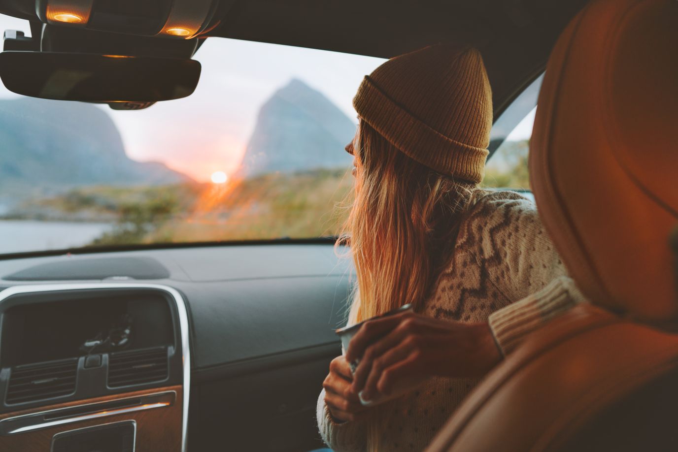 en jente som sitter i bilen og drikker kaffe, med solnedgang over fjellene i bakgrunnen