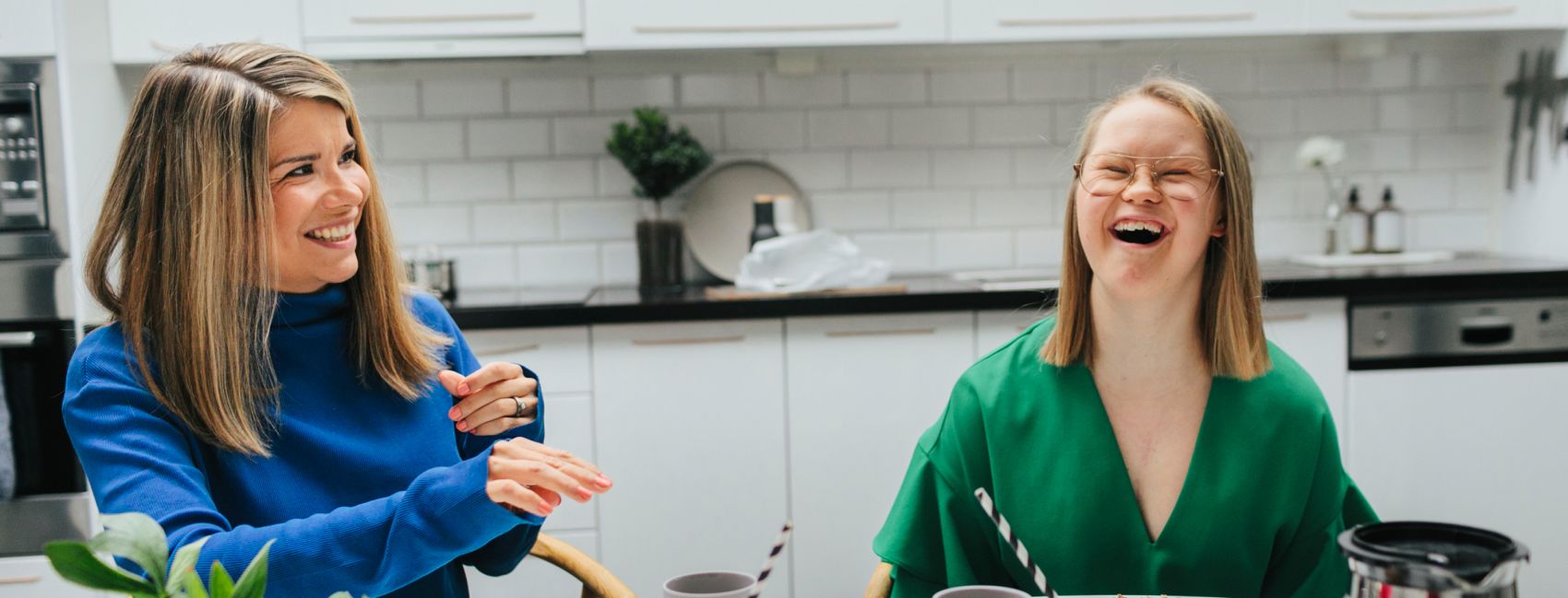 En voksen kvinne sitter ved kjøkkenbordet, ved siden av henne sitter det en jente med downsyndrom, hun smiler.