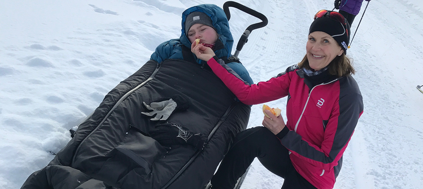Funksjonhemmed ung jente ligger i en pulk i skisporet, ved siden av sitter moren.