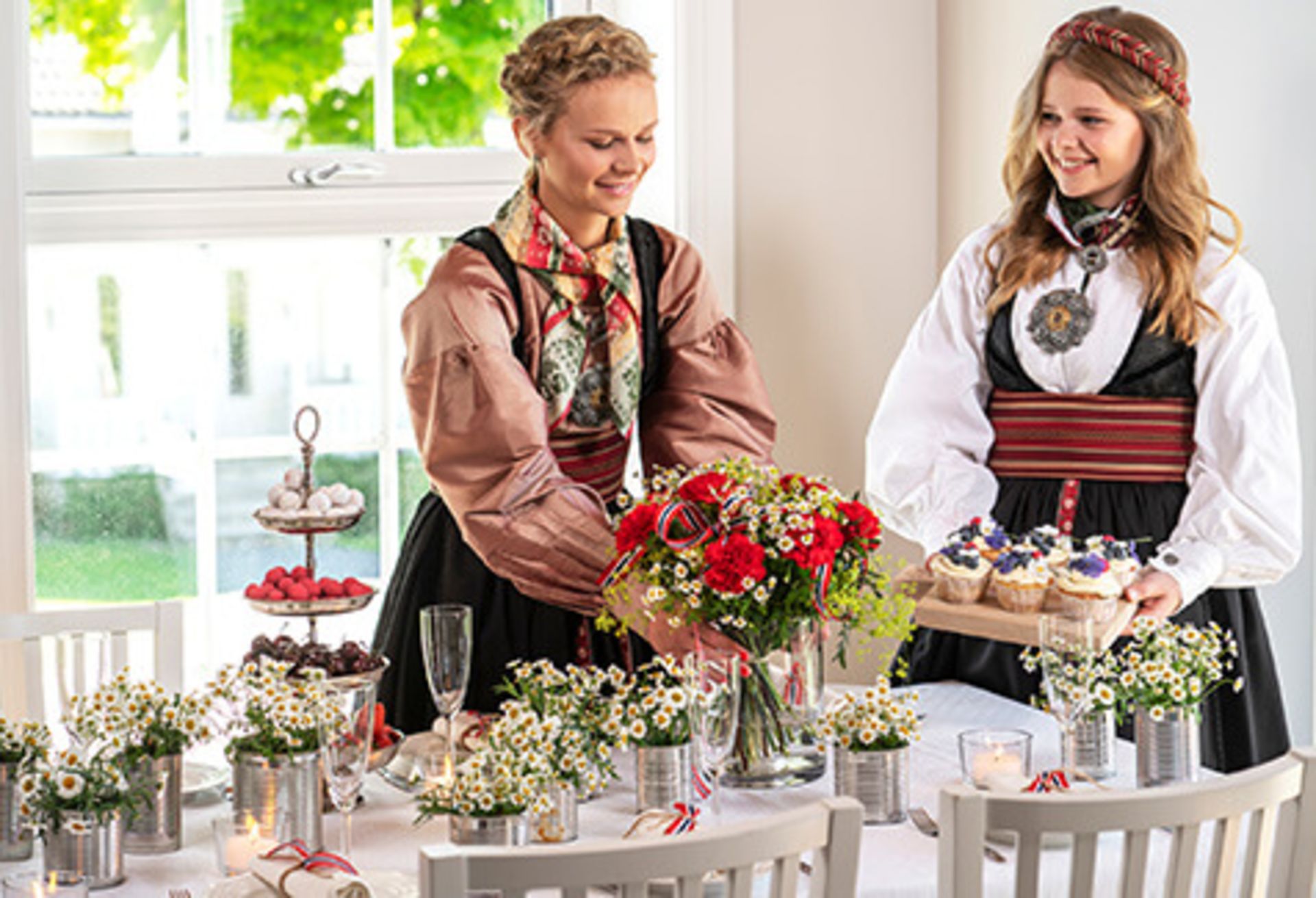 To kvinner i bunad og festdrakt pynter til 17. mai
