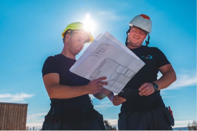 To elektrikere ser på plantegninger og smiler. Sol og blå himmel i bakgrunnen