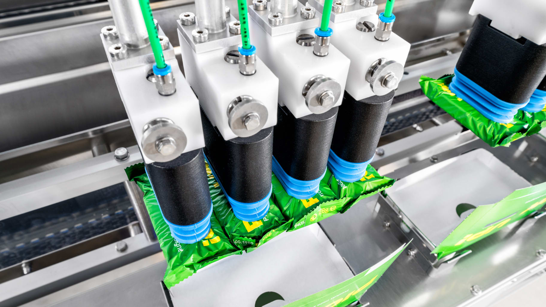 Ein Robotersauggreifer, der vier in Schlauchbeutel verpackte Keksriegel hält. Direkt darunter befinden sich leere, aufgerichtete Kartons.