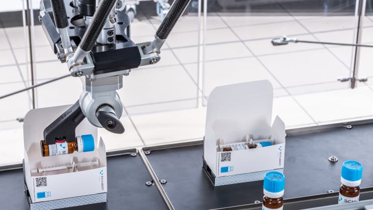 In a packaging line from Schubert-Pharma, a robot places a glass vial with other pharmaceuticals in the erected display box.