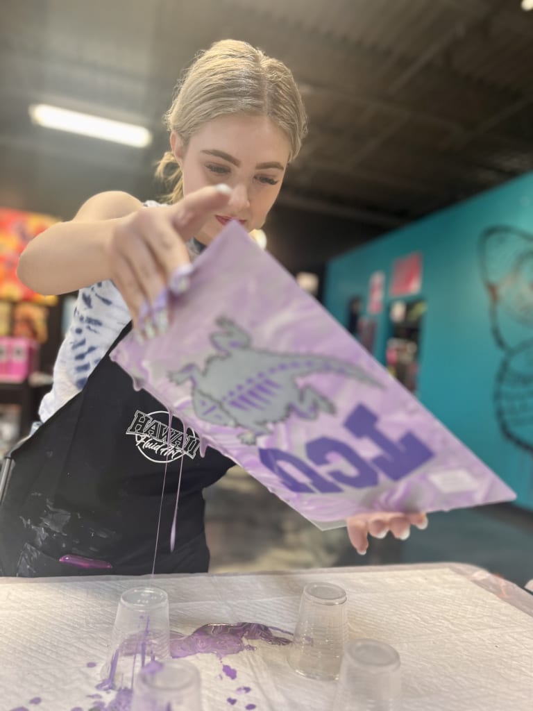 Guest dripping purple paint from a dragon-stencil canvas during a fluid art pour at ATL Fluid Art in Brookhaven, Atlanta