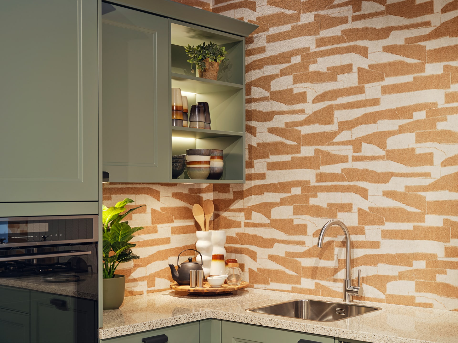 Een moderne L-keuken van Kitchen4All in de stijl Landelijk rust. De keuken heeft matte groentinten fronten en een grijs composiet werkblad. De achterwand is voorzien van een opvallend patroon in aardetinten.