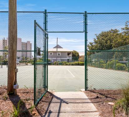 Tennis court with fenced perimeter surrounded by mature landscaping and residential buildings.