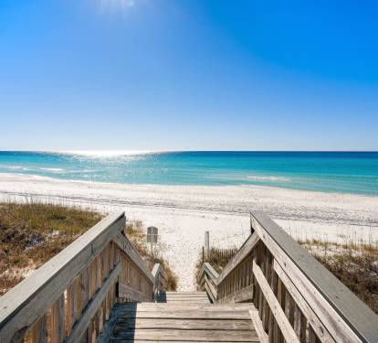 Wooden boardwalk leads to pristine white sand beach with crystal-clear turquoise waters stretching to the horizon.