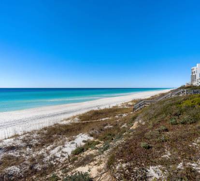 Pristine white sand beach stretches along turquoise waters under brilliant blue skies, with coastal properties nearby.