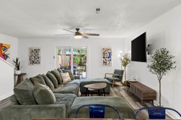 Settle into Unit A's oversized green sectional for a movie on the large smart TV. High-speed Wi-Fi, colorful abstract art, and an open kitchen/dining layout make it a welcoming gathering space. Sliding doors lead to the covered patio and yard