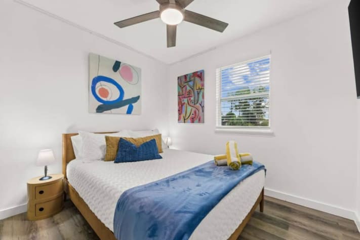 Unit B's upstairs queen bedroom sleeps two and has its own smart TV, bedside lighting, a ceiling fan, and colorful abstract art. Fresh bed linens and towels are provided. It shares the hallway bathroom, with a bathtub and shower, with the bunk room