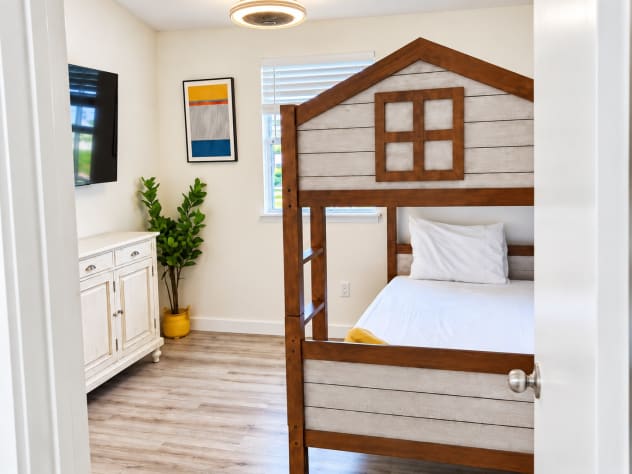 Unit A's upstairs bunk room features a playful house-frame bunk bed, smart TV, a storage cabinet, and colorful wall hooks. Bed linens and towels are provided. The room shares the hallway bathroom, with a bathtub and shower, with the queen room