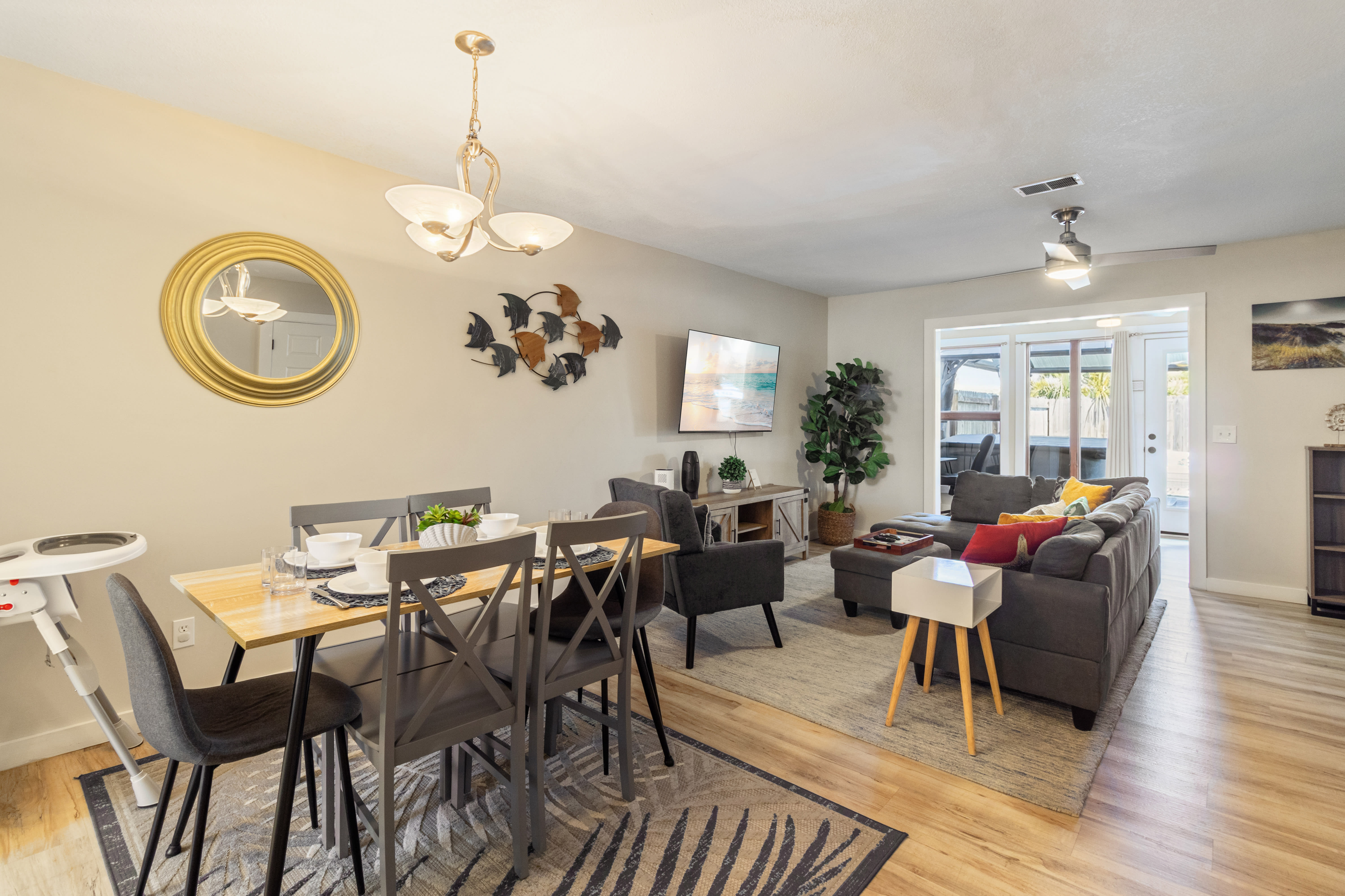 Bright open-concept living and dining space with coastal d&eacute;cor, featuring a dining table set for guests, a comfortable sectional sofa, wall-mounted TV, and stylish beach-themed accents. Warm and welcoming layout ideal for relaxing or entertaining.