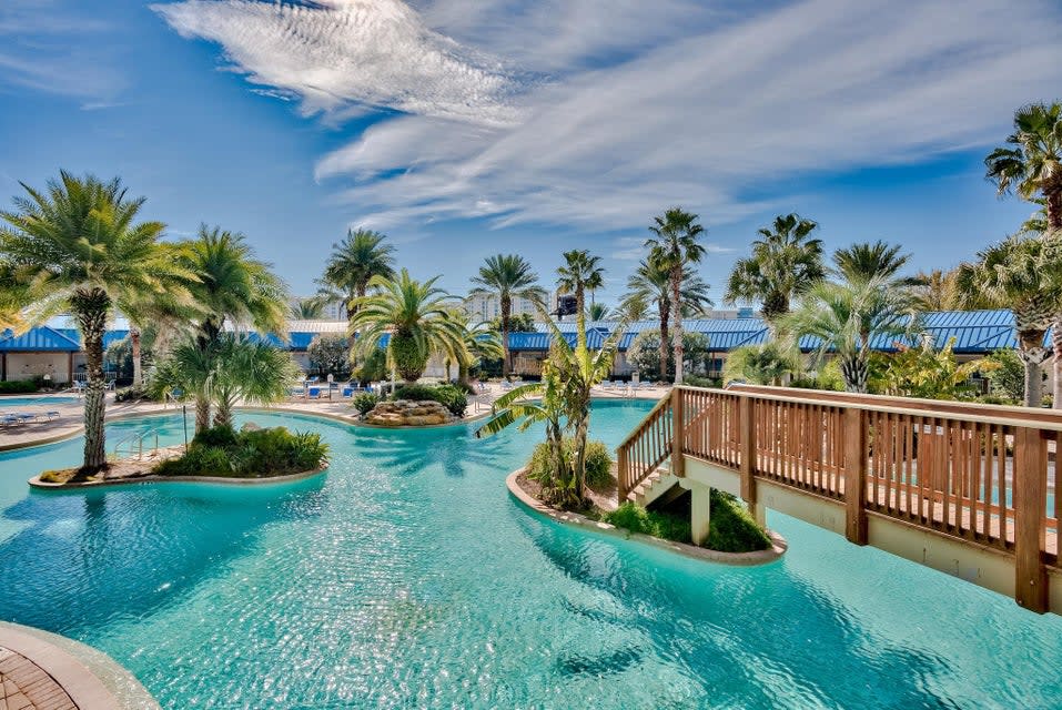 Resort-style pool w/ winding waterways, palm trees, and a sunny bridge connecting lush islands.