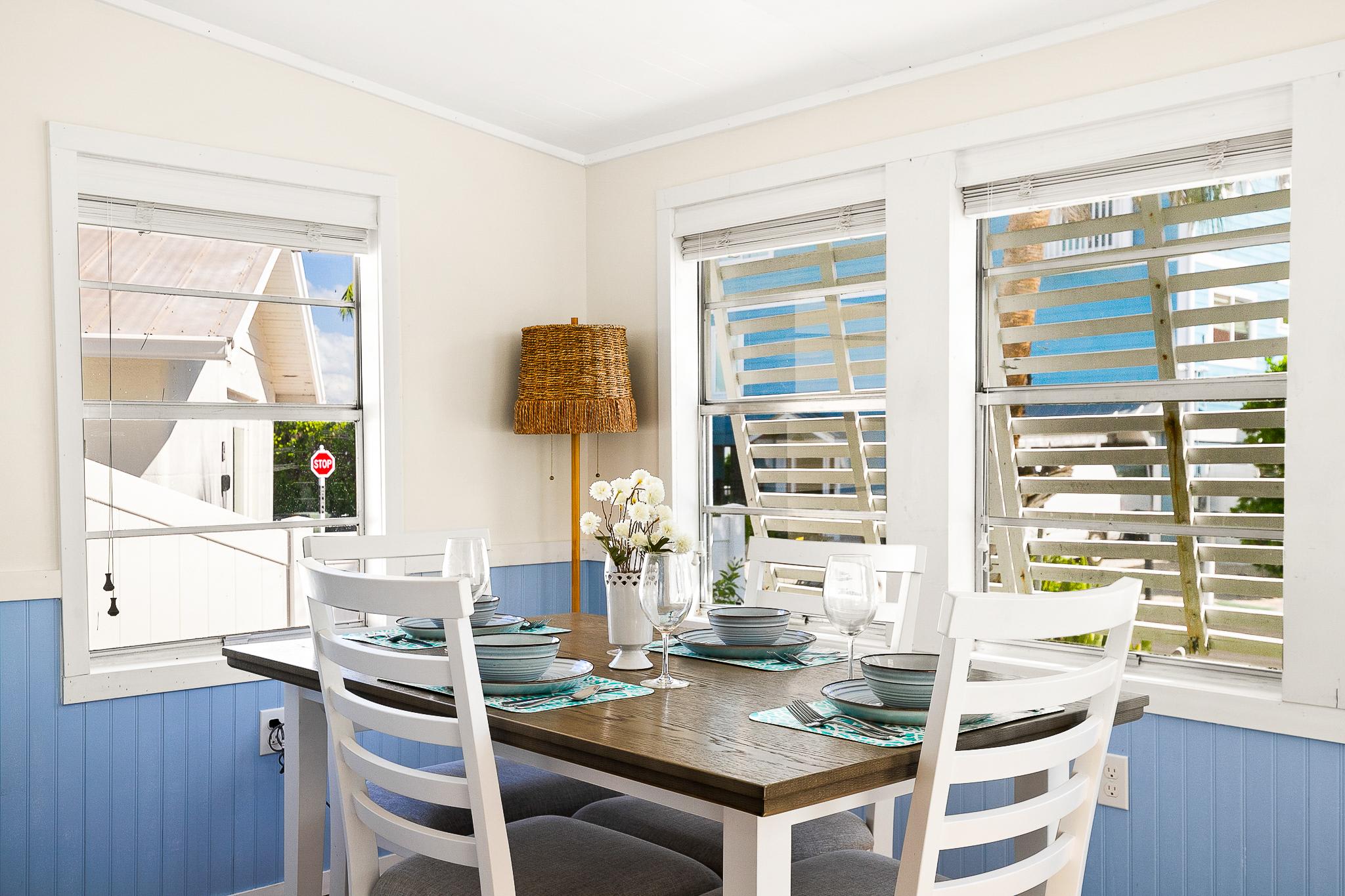 This bright dining nook features large windows, coastal blue accents, and a wooden table set for four—perfect for casual meals in a beach cottage setting.