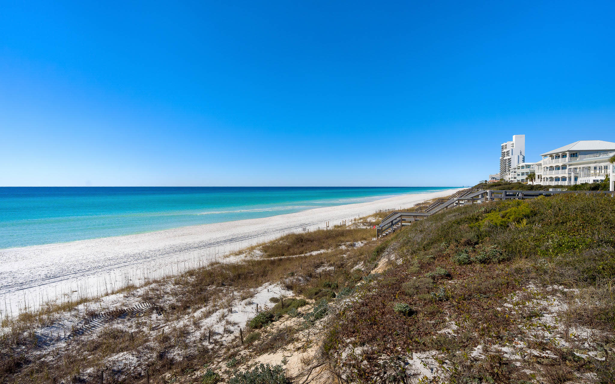 Pristine white sand beach stretches along turquoise waters under brilliant blue skies, with coastal properties nearby.