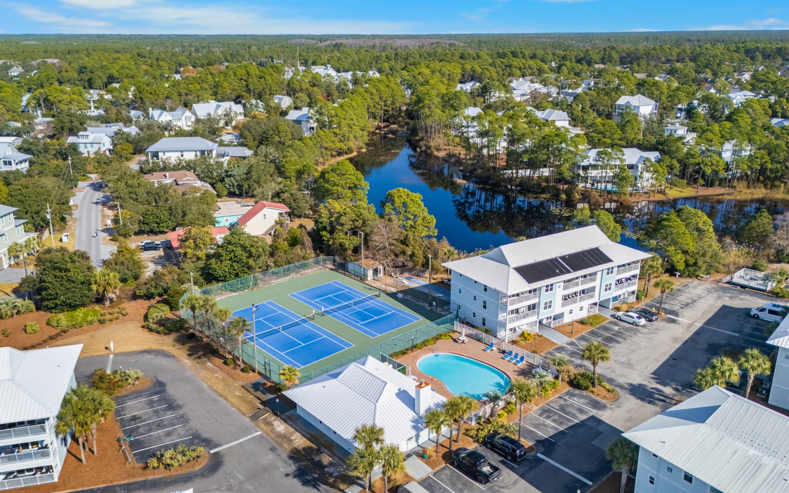 Aerial view of a waterfront community featuring tennis courts, swimming pool, and beautiful lake surrounded by lush trees and residential homes.
