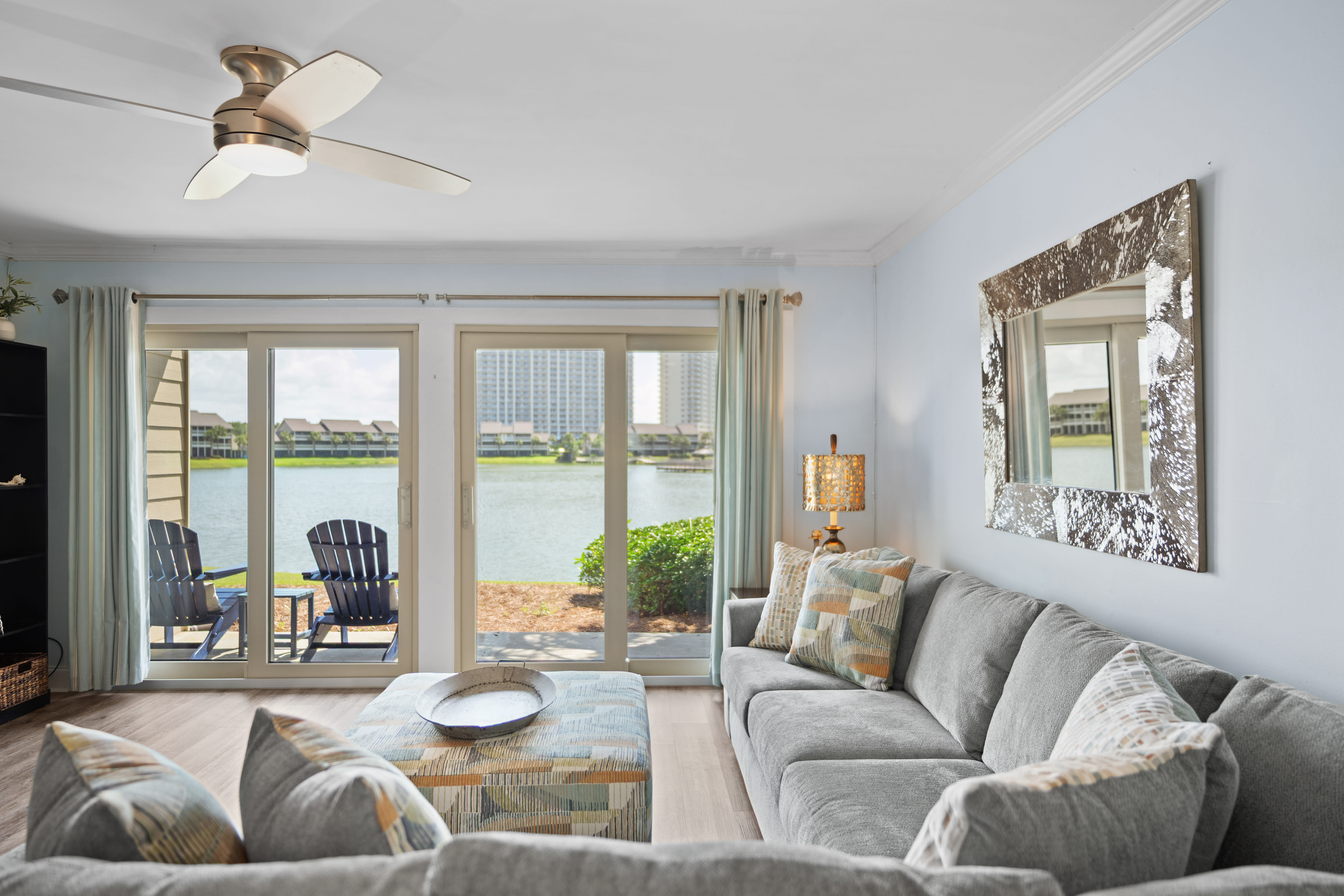 Coastal living room with plush sectional, patterned ottoman, and large glass doors opening to a lakefront patio. Light-filled and serene with stylish, beachy accents.