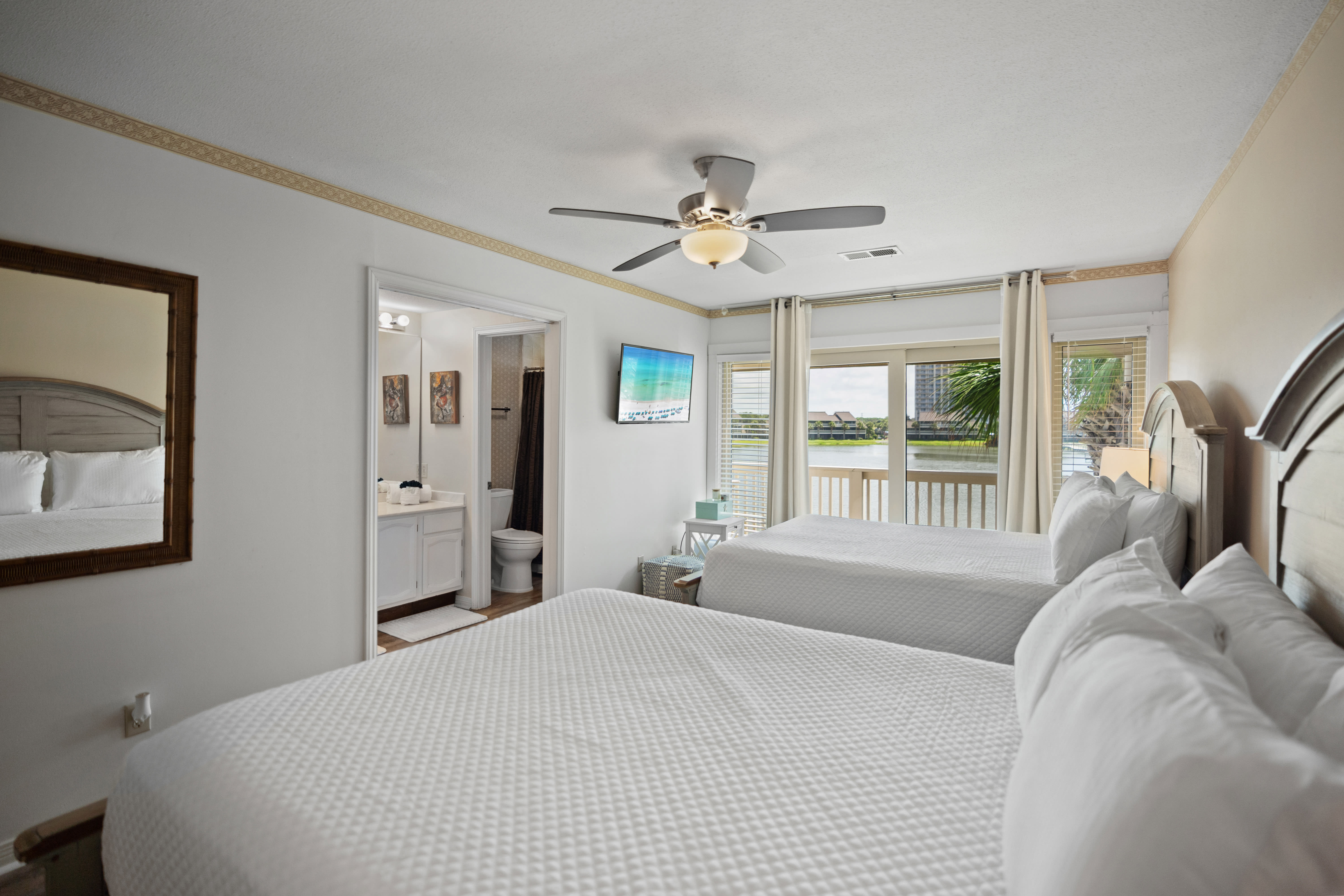 Lakeview guest bedroom with two queen beds, ensuite bathroom, wall-mounted TV, and sliding doors to a private balcony. Features white bedding, soft natural light, and a ceiling fan for comfort.