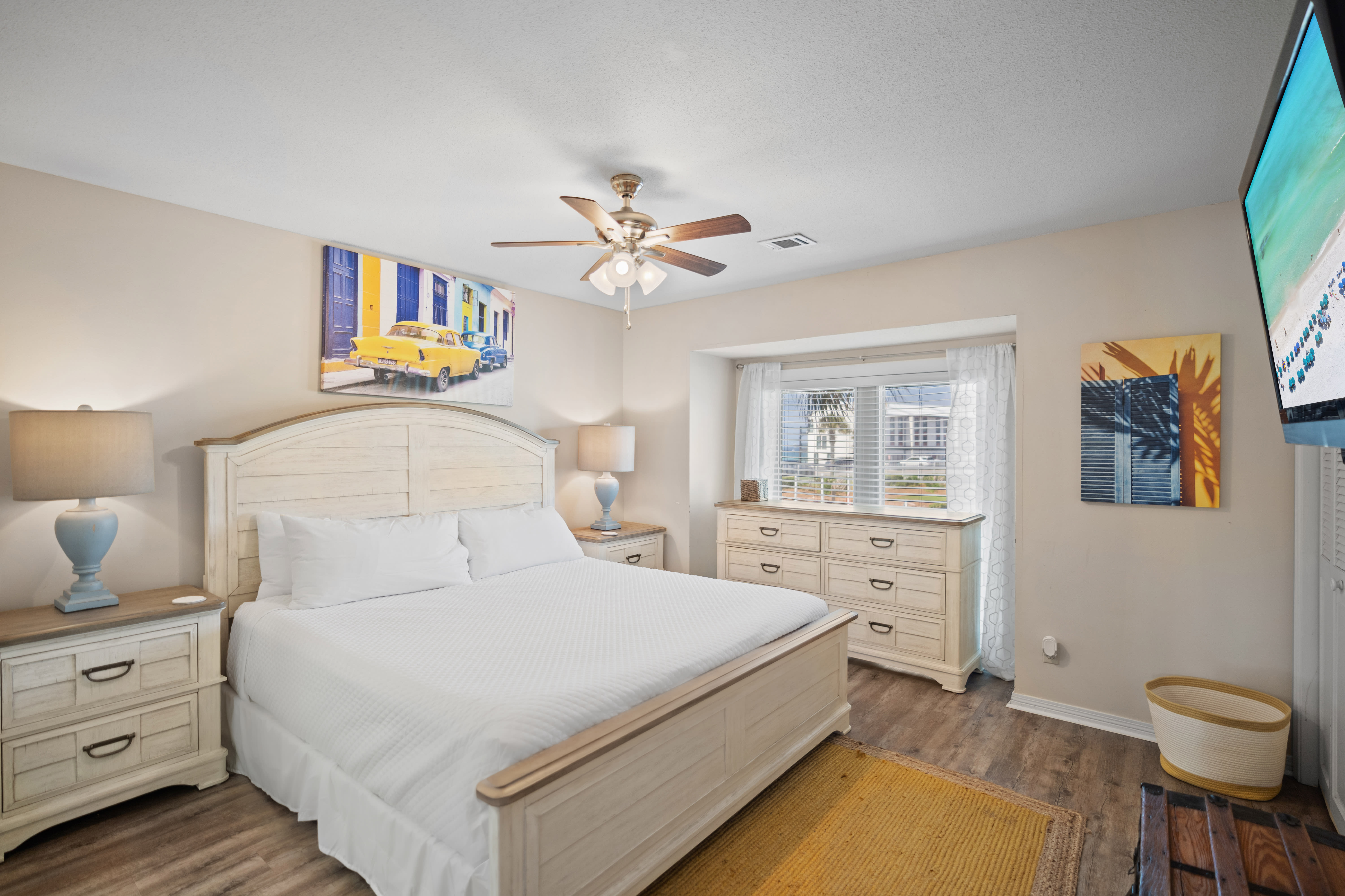 Inviting primary bedroom with a king bed, light wood furniture, and bright window nook. Features soft neutral tones, wood-look flooring, a ceiling fan, and vibrant wall art for a cozy, beachy feel.