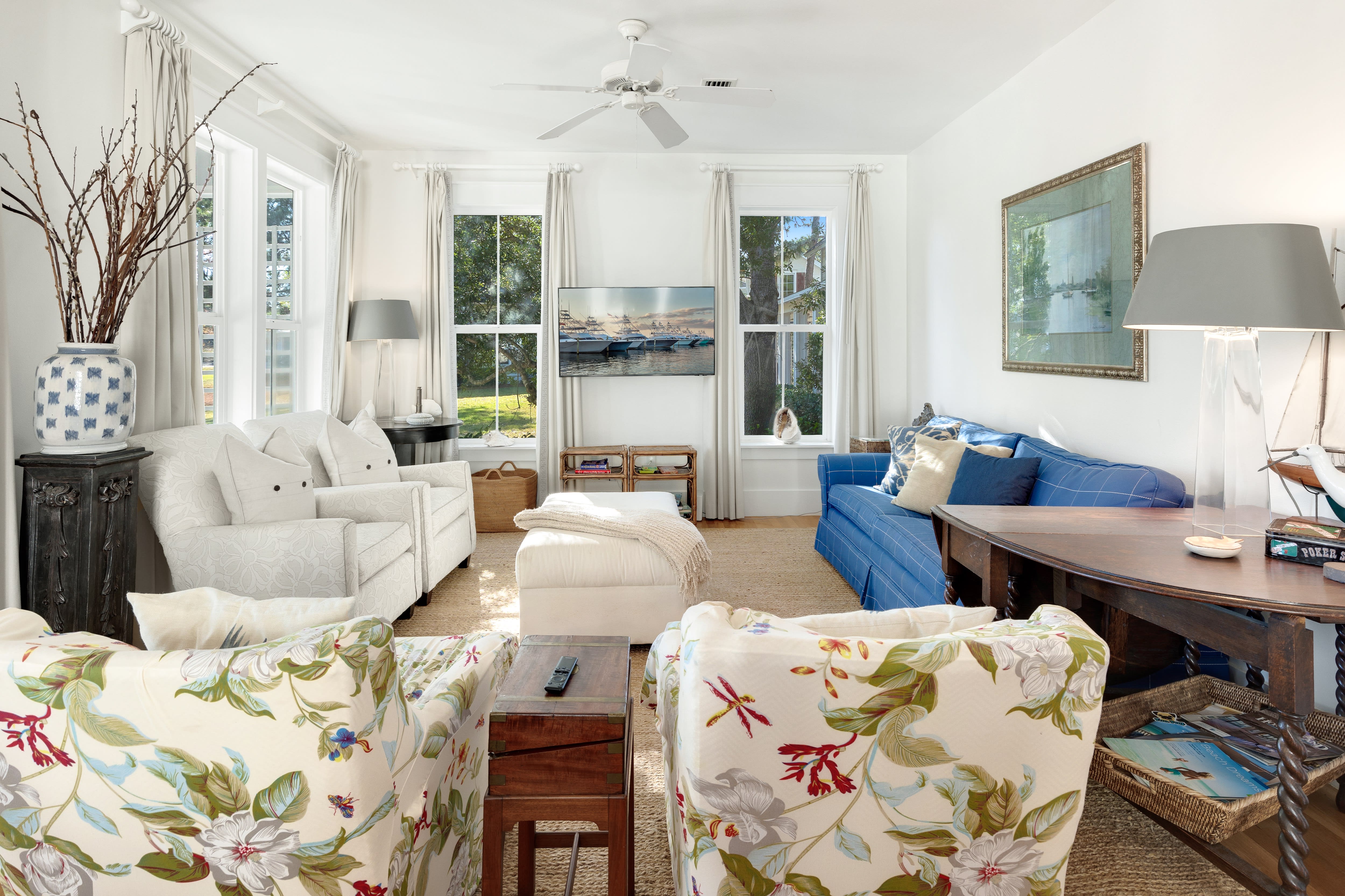 Your bright living room invites relaxation with cozy seating, coastal artwork, and natural light streaming through windows overlooking lush greenery.