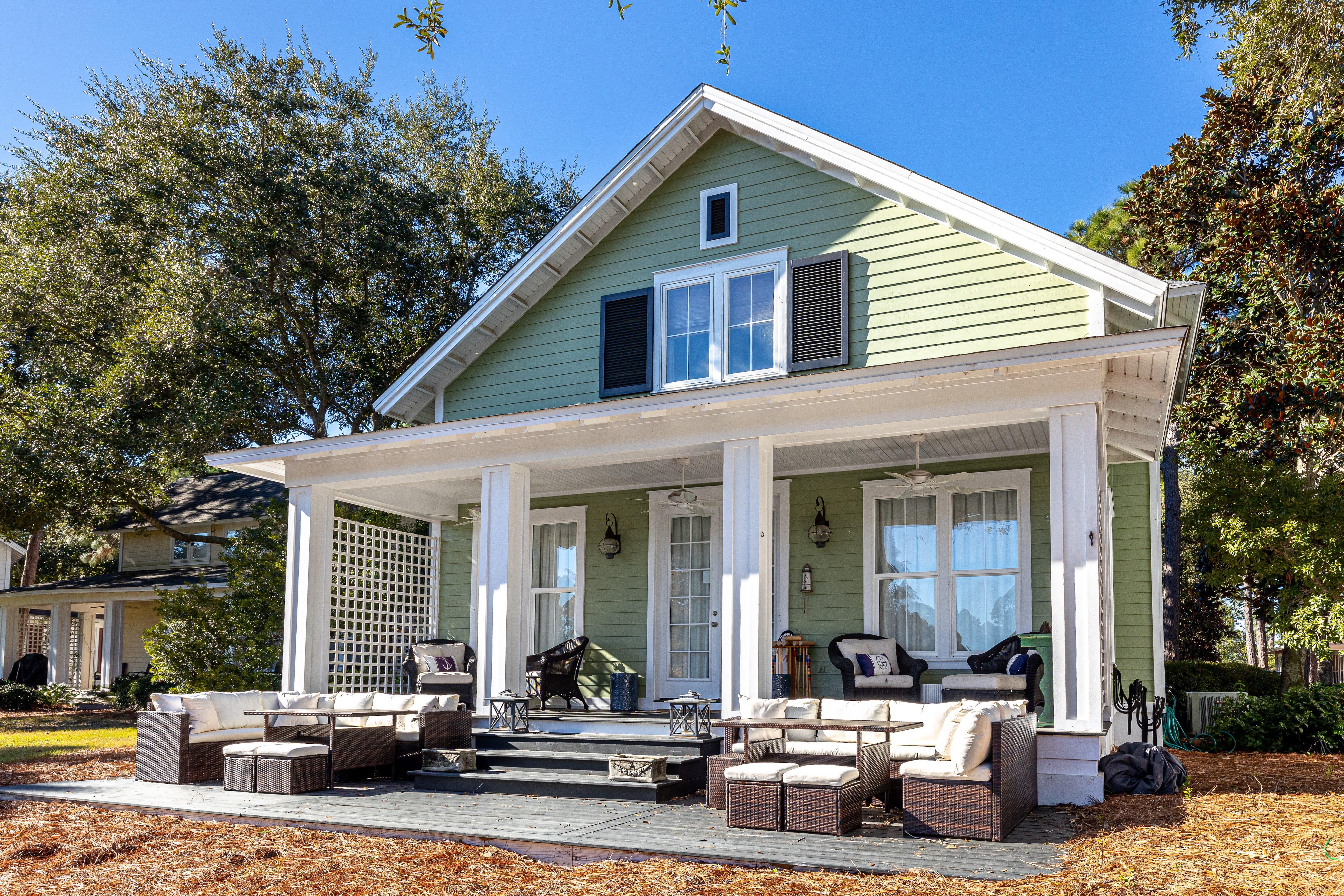 Charming green cottage with spacious wraparound porch featuring comfortable outdoor seating surrounded by mature trees on sunny day.