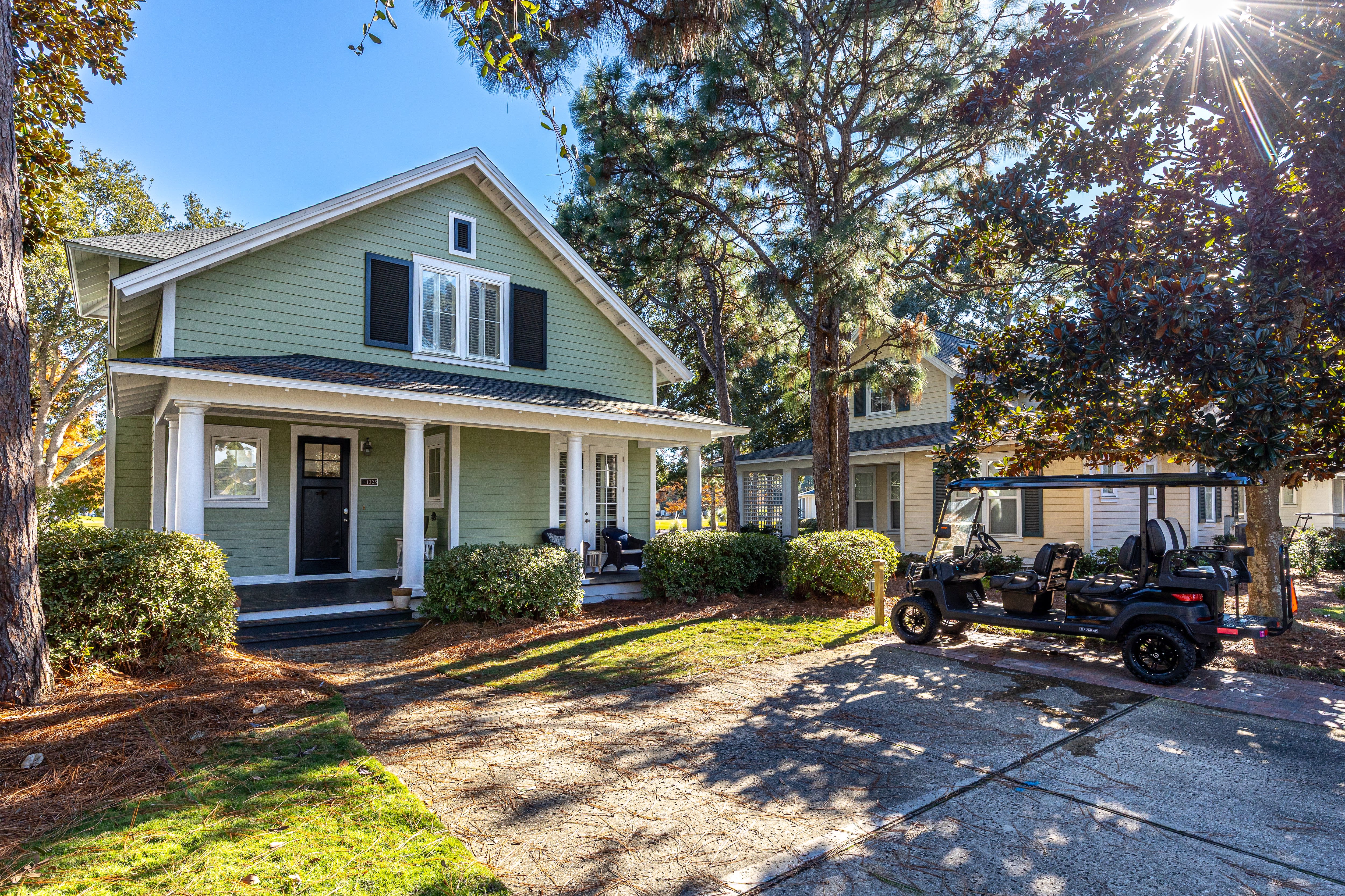 Charming green cottage with front porch and convenient parking area, nestled among mature trees in a peaceful neighborhood setting.